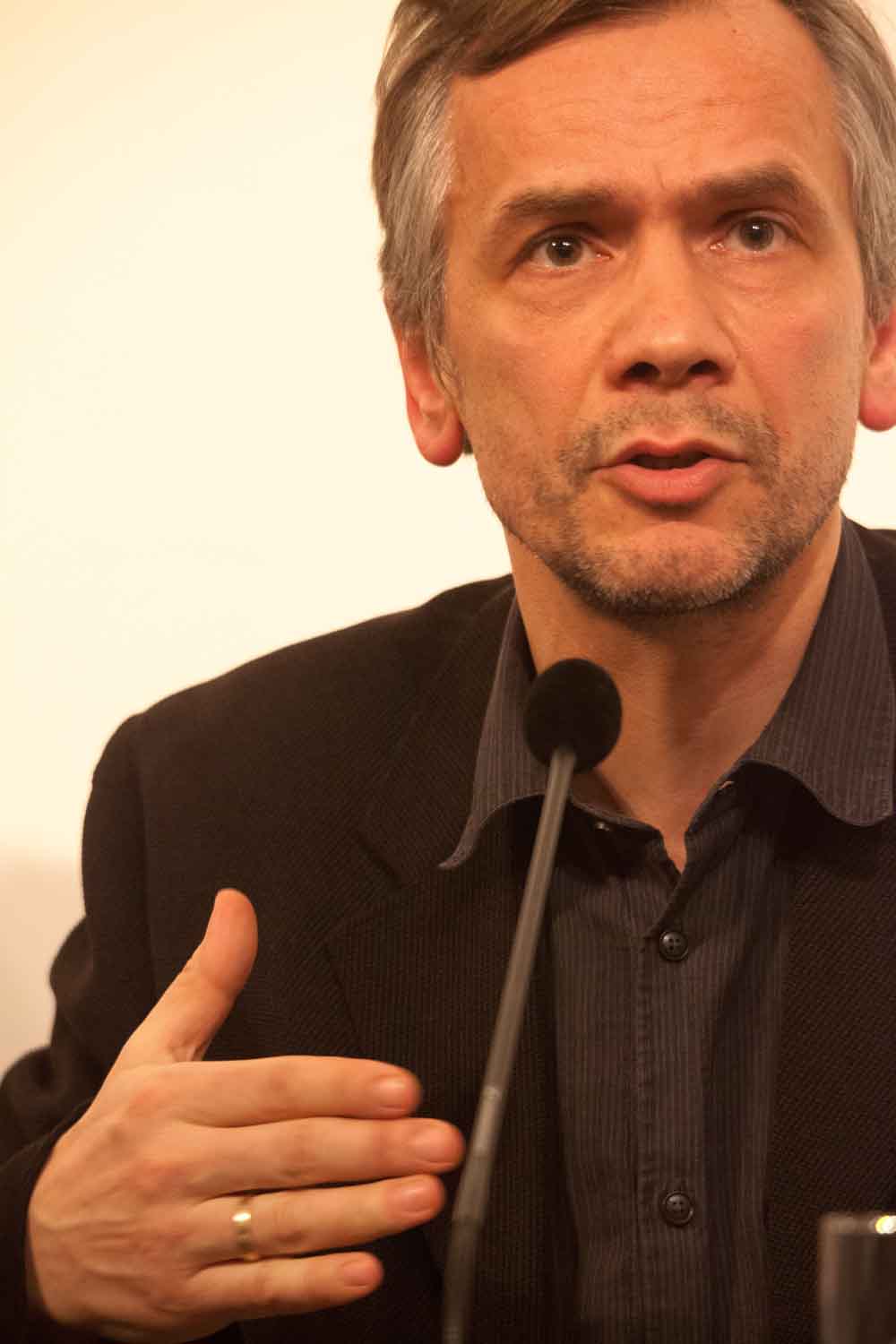 Zielinski.Photography - Images of artworks and culture - Berlin Zielinski, wearing a black suit, is speaking into a microphone during his music performance.