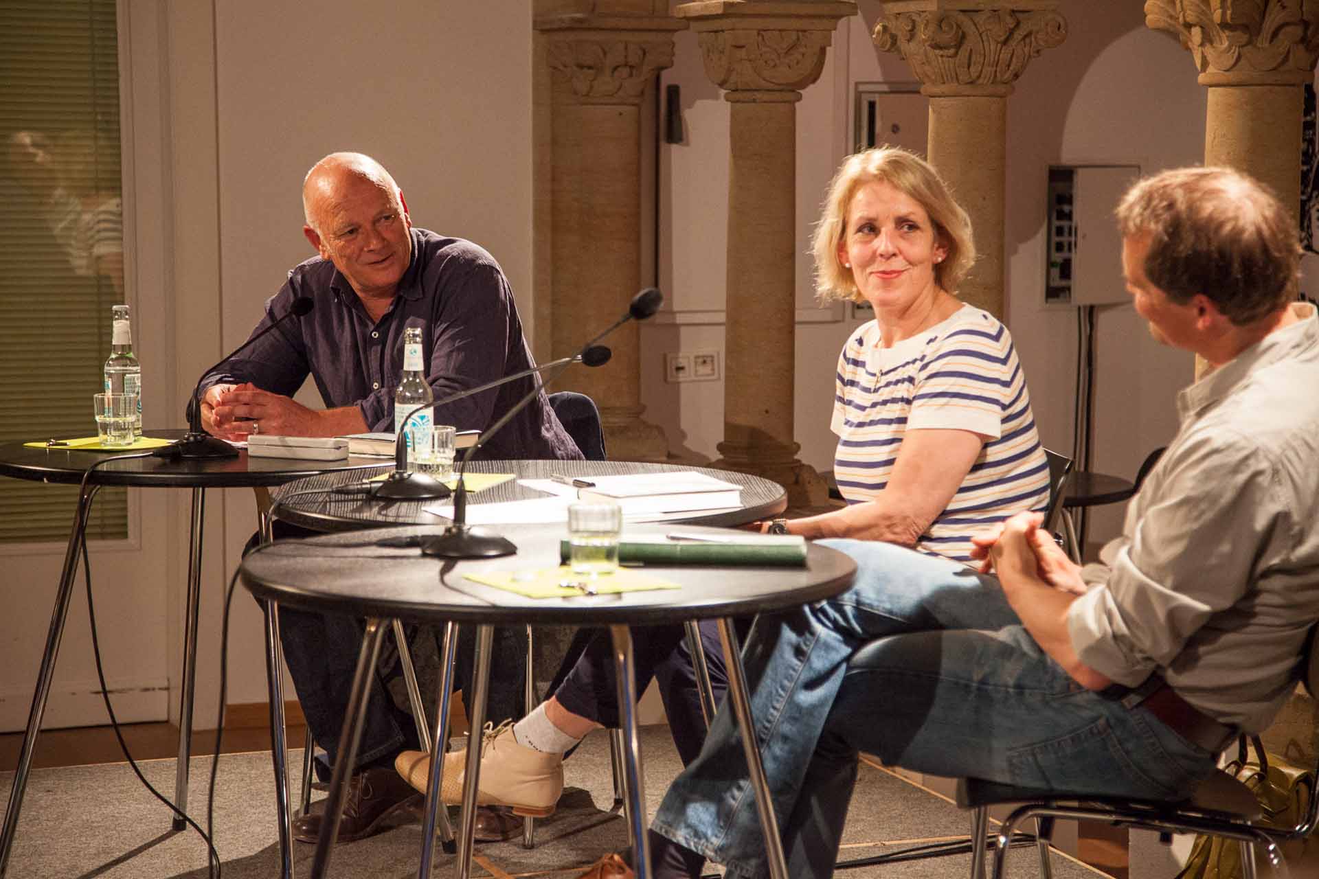 Zielinski.Photography - Images of artworks and culture - Berlin Three people engaged in a performance, seated at a table in front of a microphone.