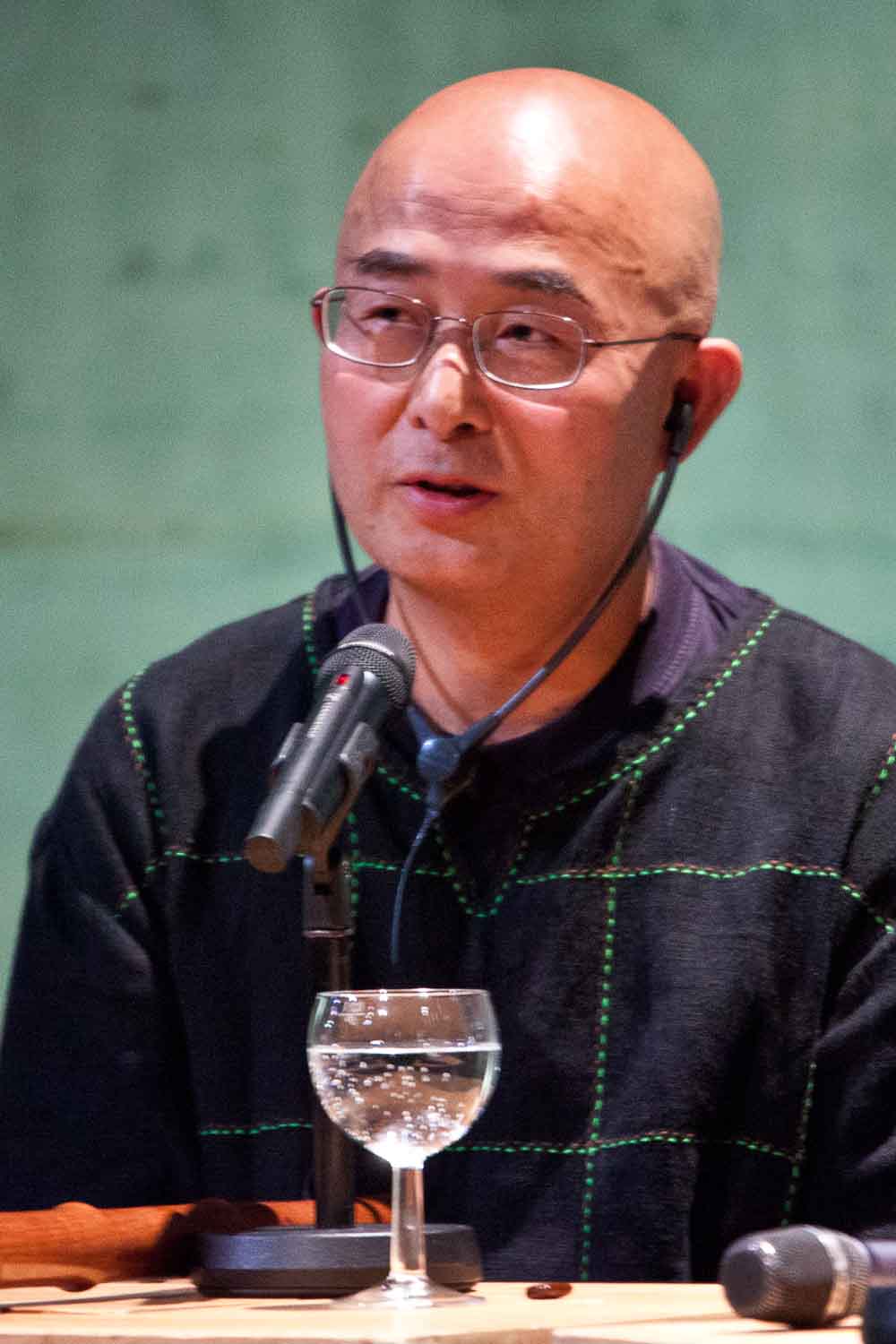 Zielinski.Photography - Images of artworks and culture - Berlin An exhibition of a bald Asian man sitting at a table with a microphone, delivering a performance.