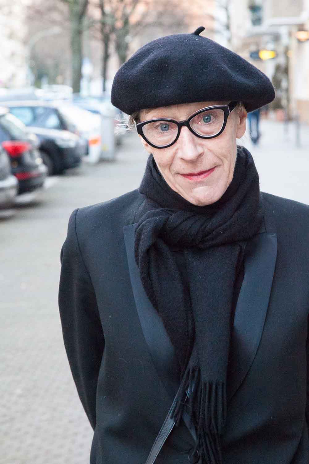 Zielinski.Photography - Images of artworks and culture - Berlin A woman wearing glasses and a beret standing on a street, captivated by the performance of vibrant artworks exhibited around her.