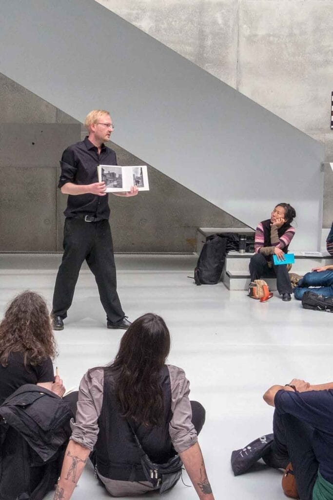 Zielinski.Photography - Gallery Exhibition Photography, Professional Art Documentation - Berlin A man is giving a performance, reading a book to a group of people.