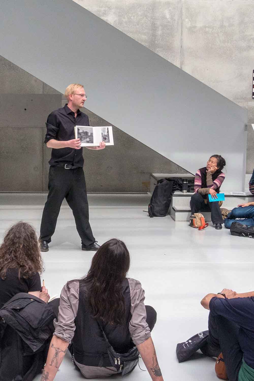 Zielinski.Photography - Images of artworks and culture - Berlin A man is giving a performance, reading a book to a group of people.
